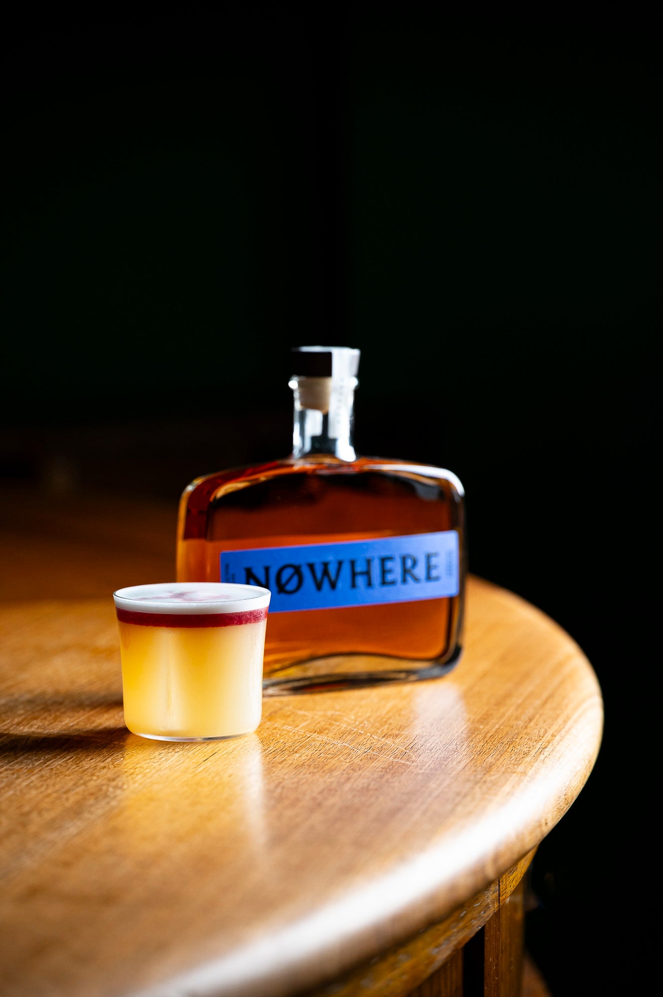 A bottle of amber liquor with a blue label reading NOWHERE stands on a wooden table beside a small clear shot glass filled with the same amber liquid.