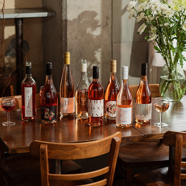 A stunning lineup of rosé wine bottles on a rustic dining table with fresh flowers — showcasing the variety in Ginny's Mystery Rosé Case.